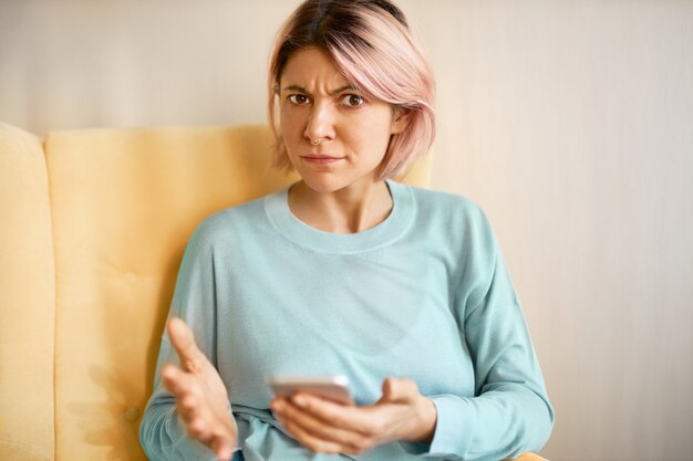 Toma en interiores de una mujer joven e irritada disgustada con una sudadera azul sentada en un sofá con un teléfono celular, que tiene problemas de conexión o carga baja de la batería, mirando a la cámara con expresión facial molesta