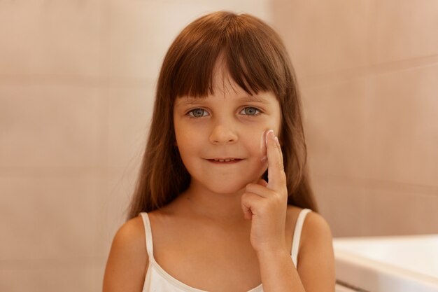 Toma en interiores de una niña con crema facial en la mejilla, señalando con el dedo en la cara, mirando directamente a la cámara, vistiendo una camiseta blanca sin mangas, realizando procedimientos de belleza sola.