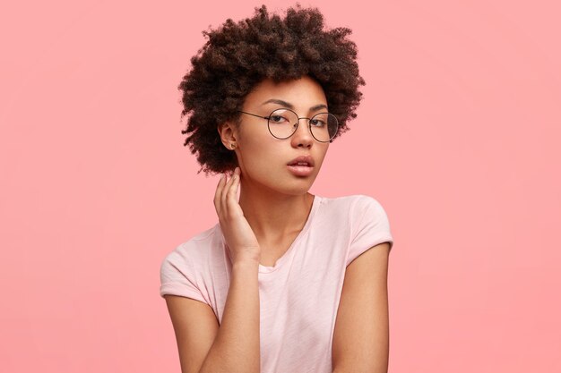 Toma lateral de una adorable joven de piel oscura, se ve seria y con confianza, muestra su belleza natural, posa para una revista de moda, usa gafas redondas y una camiseta informal