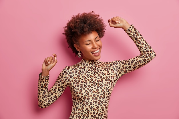 Toma de medio cuerpo de una mujer alegre y complacida con cabello afro, baila con los brazos en alto, disfruta de la vida, inspirada durante la sesión de fotos