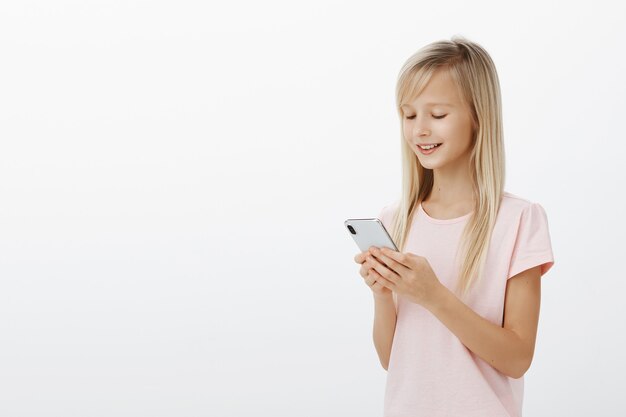 Toma de perfil medio vuelta de una niña creativa inteligente con cabello rubio en una camiseta rosa, sosteniendo un teléfono inteligente y sonriendo a la pantalla, jugando un juego divertido en el dispositivo, disfrutando de pasar tiempo sobre una pared gris