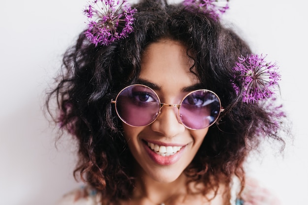 Foto gratuita toma de primer plano de alegre niña africana con flores en el pelo sonriendo. retrato de hermosa modelo de mujer negra con expresión de cara feliz.