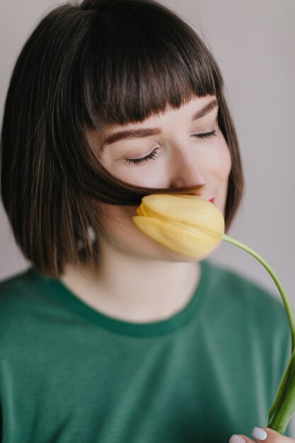 Toma de primer plano de elegante chica europea disfrutando de sabor a tulipán con los ojos cerrados. Retrato de mujer joven con corte de pelo corto con flor amarilla cerca de la cara.