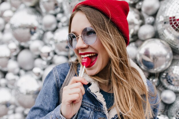 Toma de primer plano de encantadora joven divirtiéndose durante la sesión de fotos con caramelo rojo. Chica atractiva en chaqueta de mezclilla lamiendo piruleta en la pared de brillo.