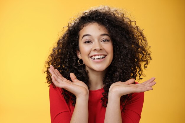 Toma de primer plano de feliz amable y tierna estudiante bastante caucásica con pelo rizado y piel perfecta sonriendo encantada sosteniendo las palmas extendidas cerca de la cara divirtiéndose sobre fondo amarillo.