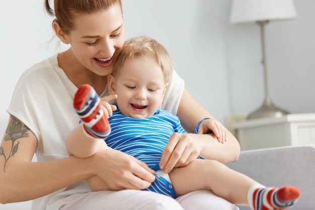 Toma de primer plano de la feliz madre y el niño en casa. Mujer joven abotonarse la ropa del niño, sosteniendo cuidadosamente al hijo en sus piernas. Niño lindo con cabello rubio en calcetines de colores mirando sus movimientos.