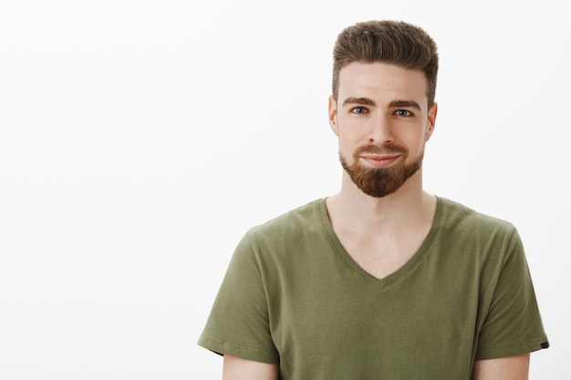 Toma de primer plano de guapo agradable caucásico barbudo con ojos azules en camiseta con cuello en v sonriendo encantado y divertido mirándote con mirada suave contra la pared blanca