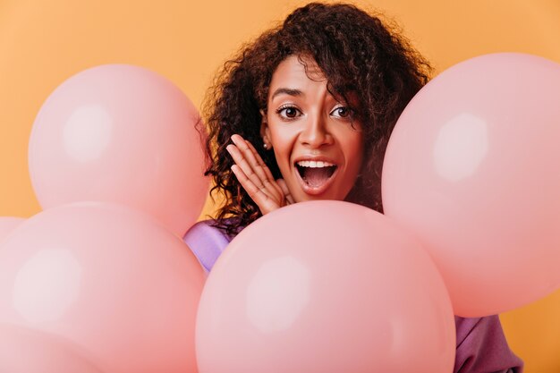 Toma de primer plano de mujer africana hermosa sorprendida aislada en naranja. Impresionante chica negra con cabello rizado celebrando un cumpleaños.