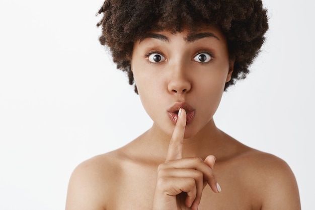 Toma de primer plano de mujer afroamericana femenina linda y tierna a punto de contar el secreto de belleza diciendo shh mientras muestra el gesto de silencio con el dedo índice sobre la boca mirando amigable