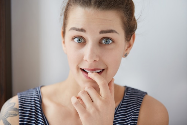 Toma de primer plano de mujer europea preocupada en top pelado en la pared blanca. Niña de ojos azules mordiendo su uña interrumpida por la ansiedad. Chica incómoda levantando cejas oscuras con miedo y sorpresa.