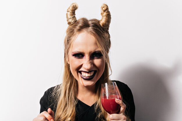 Toma de primer plano de niña alegre bebiendo sangre en halloween. Vampiro despreocupado con cabello rubio posando en el carnaval.