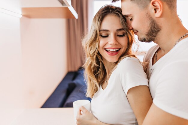 Toma de primer plano de niña de buen humor con taza de té sonriendo al marido. Foto interior de mujer rubia rizada disfrutando de un café con su novio.