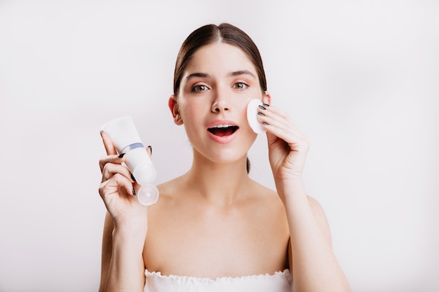 Toma de primer plano de niña de ojos verdes haciendo procedimiento de spa en la pared blanca. Mujer humedece su rostro con crema Dove.