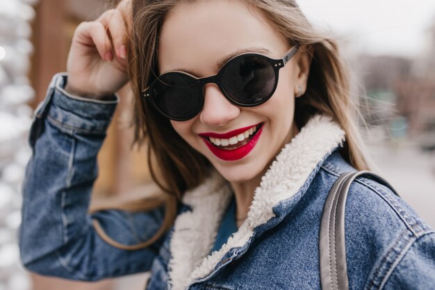 Toma de primer plano de la romántica chica caucásica posando con una hermosa sonrisa. Retrato al aire libre de dama con cabello oscuro caminando por la ciudad en fin de semana de primavera.