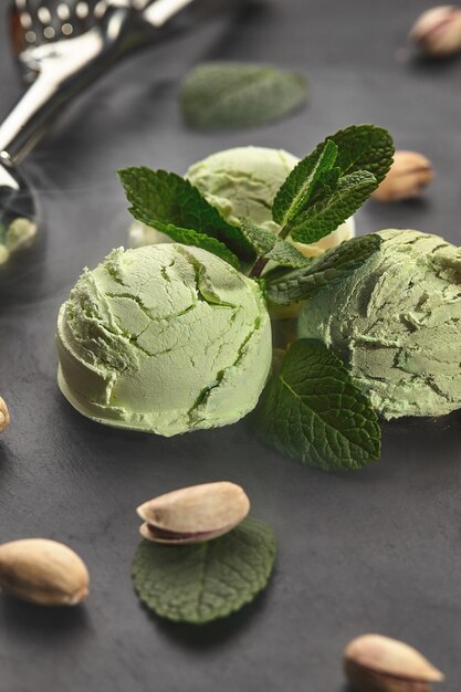 Toma de primer plano de un sabroso helado de pistacho decorado con menta, hay pistachos dispersos cerca, servidos con una cuchara de metal en una pizarra sobre un fondo negro.