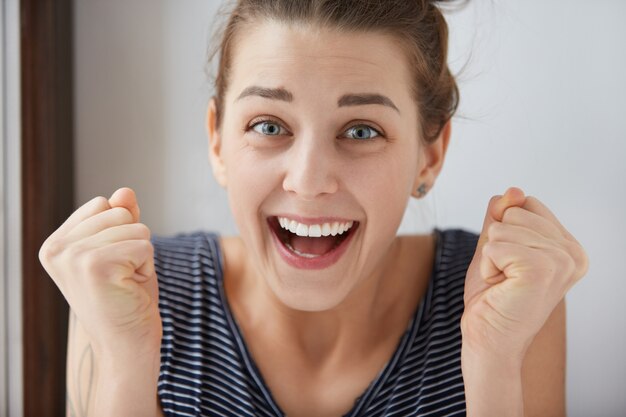 Toma de primer plano de sonriente niña europea de ojos azules apretando los puños con alegría sin fin. Las emociones positivas y la sensación de merecido triunfo hacen que la mujer encantadora se vea hermosa en la pared blanca.