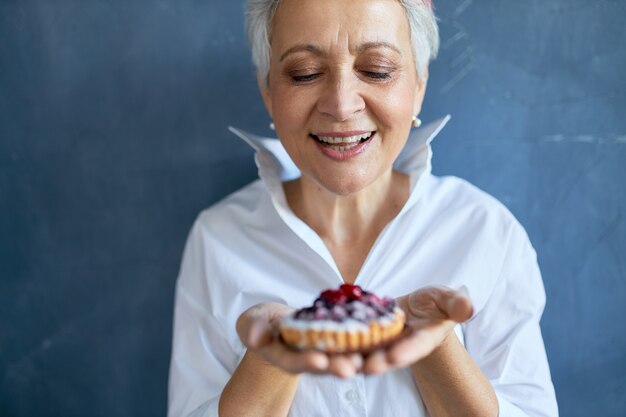 Toma recortada de abuela atractiva alegre en camisa blanca sosteniendo un trozo de pastel de bayas recién horneado para cumpleaños, con expresión facial alegre, sonriendo ampliamente.