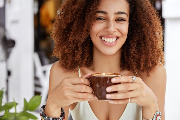 Toma recortada de atractiva mujer afroamericana alegre sostiene una taza de café, almuerza en una acogedora cafetería, descansa después de la excursión, recrea en el país de vacaciones