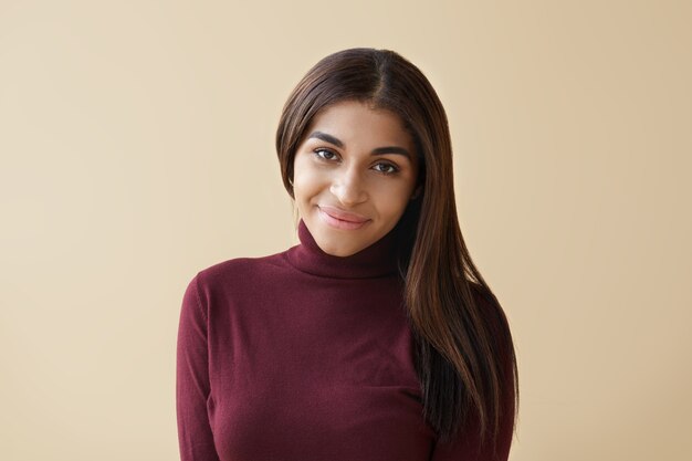 Toma recortada de hermosa joven mestiza con cabello largo oscuro sonriendo felizmente mientras se regocija con las buenas noticias. Atractiva mujer africana de buen humor, expresando alegría y emociones positivas