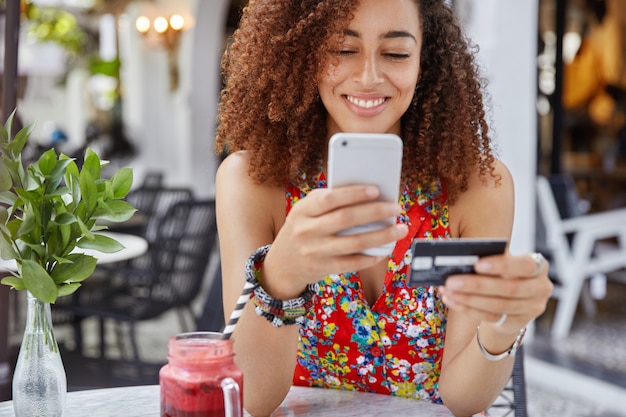 Toma recortada de hermosa mujer afroamericana con mirada feliz, tiene teléfono celular moderno y tarjeta de crédito, hace compras en línea