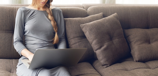 Toma recortada de joven rubia con trenza trabajando en una computadora portátil sentada en el cómodo sofá oscuro en casa, luz cálida retroiluminada. Concepto de freelance o estilo de vida.