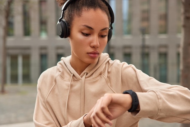 Foto gratuita toma recortada de mujer deportiva usa reloj inteligente al aire libre, verifica las calorías quemadas, escucha música a través de auriculares vestida con una sudadera beige