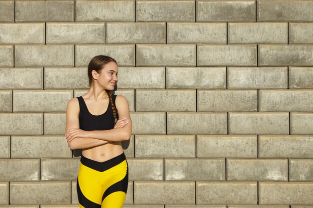 Toma recortada de mujer joven hermosa positiva con ropa deportiva negra y amarilla de moda que descansa al aire libre, posando contra la pared de ladrillo en blanco con espacio de copia para su contenido