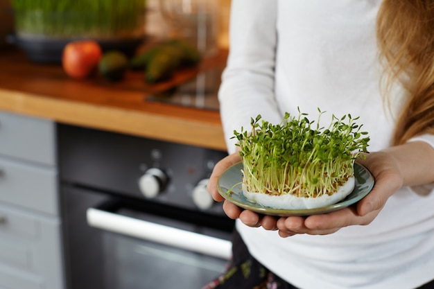 Toma recortada de una mujer sosteniendo en sus manos un platillo con un micro verde de brotes orgánicos cultivados en casa contra el acogedor interior de la cocina. Concepto de alimentos crudos saludables. Copie el espacio para el texto. Enfoque selectivo