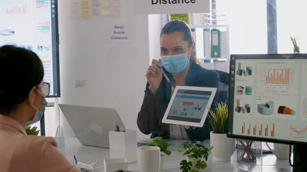 Toma trasera de mujeres de negocios con mascarilla médica trabajando juntas en la presentación de la gerencia usando una tableta mientras están sentadas en la oficina de la empresa. Equipo respetando la distancia social