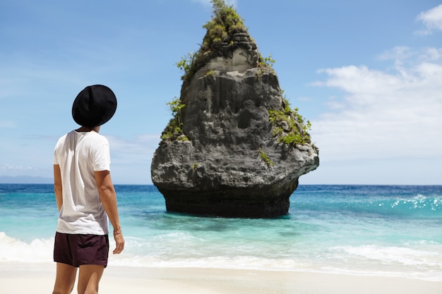 Toma trasera recortada del modelo masculino de moda con sombrero negro, camiseta y pantalones cortos de pie sobre la arena frente a un acantilado rocoso en medio del océano mientras posa en la playa