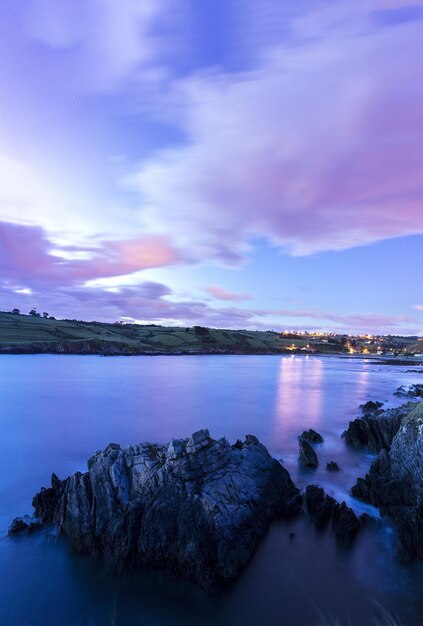 Toma vertical de un lago con rocas y una ciudad en el horizonte durante el crepúsculo Fondo de pantalla perfecto