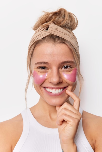 La toma vertical de una mujer positiva mantiene el dedo cerca de la boca y aplica parches de hidrogel rosa para reducir las arrugas. Lleva una camiseta informal con banda para la cabeza aislada sobre fondo blanco. Concepto de tiempo de belleza.