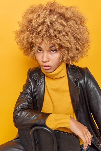 Toma vertical de poses serias de mujer de cabello rizado en la silla con mirada directa a la cámara, usa un cómodo puente con cuello y chaqueta de cuero aislado sobre fondo amarillo siendo estricto seguro de sí mismo