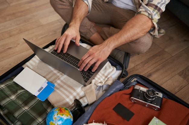 Foto gratuita tomada de primer plano de un hombre con las manos de un hombre usando una computadora portátil en la parte superior de una maleta abierta empacando para unas vacaciones