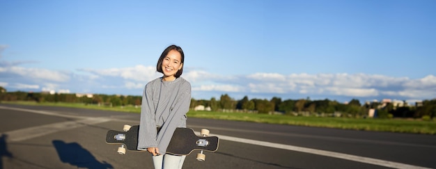 Foto gratuita tomada vertical de una chica patinadora posando con un longboard navegando en una carretera vacía en los suburbios sonriendo asiática