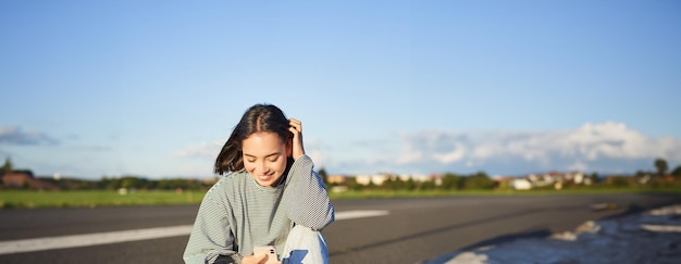 Foto gratuita tomada vertical de una mujer asiática sentada en una patineta en la carretera sosteniendo una aplicación de teléfono inteligente patinadora patines para niñas