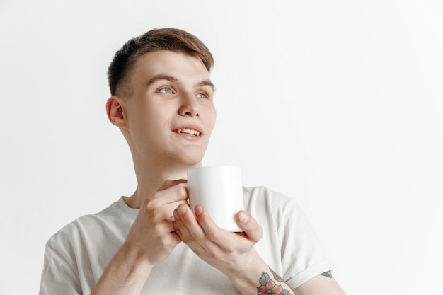 Tomando un café. Apuesto joven sosteniendo la taza de café, sonriendo mientras está de pie contra el fondo gris del estudio