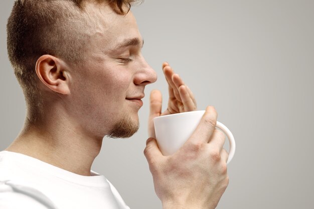 Tomando un café. Apuesto joven sosteniendo la taza de café, sonriendo mientras está de pie contra la pared gris