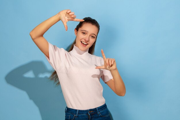 Tomando una selfie, encuadrando. Retrato de la muchacha adolescente caucásica sobre fondo azul. Preciosa modelo en ropa casual. Concepto de emociones humanas, expresión facial, ventas, publicidad. Copyspace. Se ve lindo, feliz.