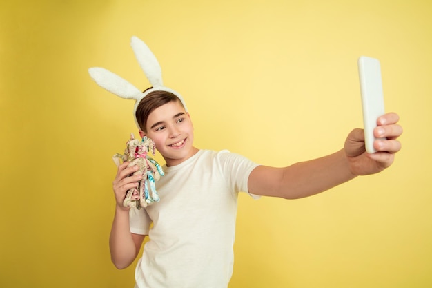 Tomando selfie con juguete. Chico caucásico como un conejito de Pascua sobre fondo amarillo de estudio. Saludos felices de pascua.