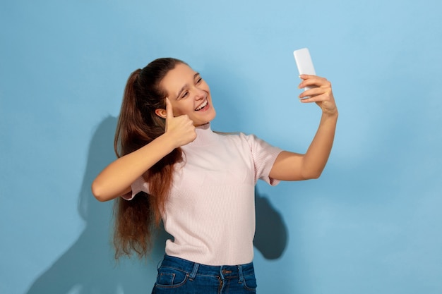 Tomando selfie o vlog, sonriendo, pulgar hacia arriba. Retrato de la muchacha adolescente caucásica sobre fondo azul. Preciosa modelo en casual. Concepto de emociones humanas, expresión facial, ventas, publicidad. Copyspace. Se ve feliz.