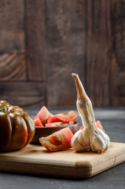 Tomate Kumato con rodajas en placa, vista lateral de ajo en pared de azulejos grises y piedra