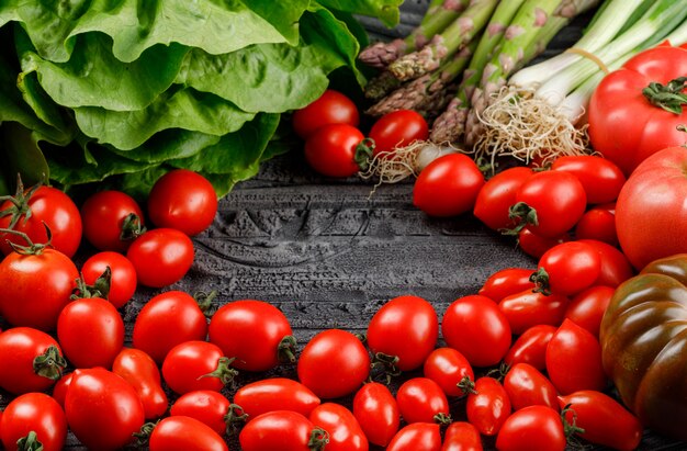 Los tomates se apilan con lechuga, espárragos, cebollas verdes en la pared de madera gris, vista de ángulo alto.