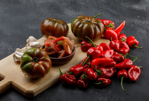 Foto gratuita tomates kumato con rodajas, pimientos rojos, bulbos de ajo en la pared gris y tabla de cortar, vista de ángulo alto.