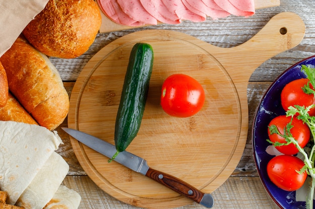 Tomates con pan, pepino, cuchillo, salchichas, verduras en un plato sobre madera y tabla de cortar, plano.