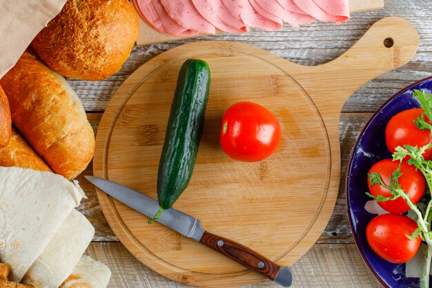 Tomates con pan, pepino, cuchillo, salchichas, verduras en un plato sobre madera y tabla de cortar, plano.