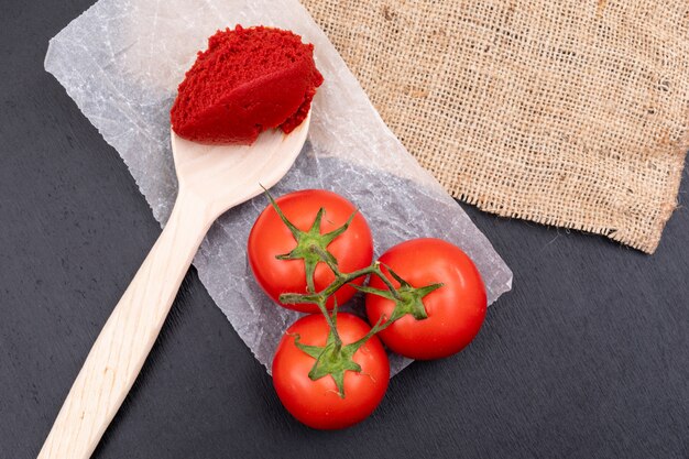 Tomates en pasta de tomate de polietileno en una cuchara de madera cerca de la tela de saco sobre superficie de piedra negra