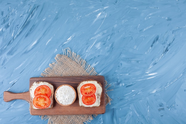 Tomates en rodajas sobre pan de queso y un tazón de harina en la tabla de cortar sobre una servilleta de arpillera en azul.