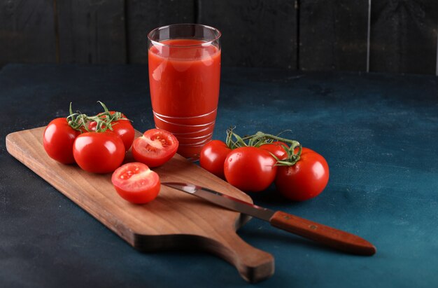 Tomates rojos y un vaso de jugo sobre una tabla de madera con un cuchillo sobre un fondo azul.