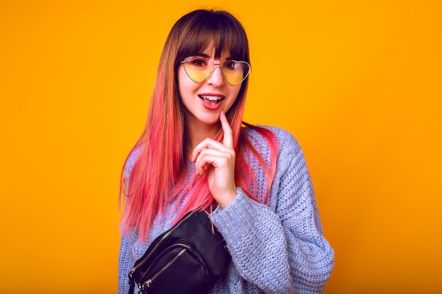 tonificado retrato de mujer feliz emocionada con inusuales pelos rosados ombre de moda posando en la pared amarilla, emociones sorprendidas, suéter acogedor y gafas de sol vintage.